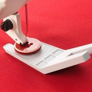 May include: Close-up of a sewing machine foot and needle, with red thread piercing a red fabric. The foot is white with a red circular base, and the fabric is a solid red color. The machine's arm is visible.