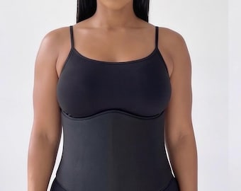 May include: A black waist trainer cincher featuring a smooth, matte finish and a structured, contoured design that sits over the midsection. The garment includes thin spaghetti straps and a fitted bodice, providing a sleek, uniform appearance.