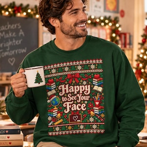 May include: A dark green crewneck sweatshirt featuring a festive graphic with the text Happy to See Your Face, surrounded by illustrations of books, apples, a globe, pencils, and a chalkboard, worn with beige pants and holding a white mug.