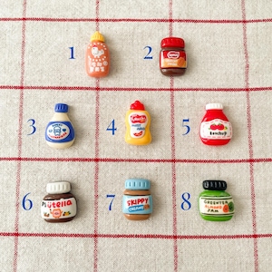 May include: Eight miniature ceramic jars and bottles with various food labels, including ketchup, mustard, peanut butter, jam, and Nutella. The jars are arranged in a grid pattern on a white and red checkered surface.