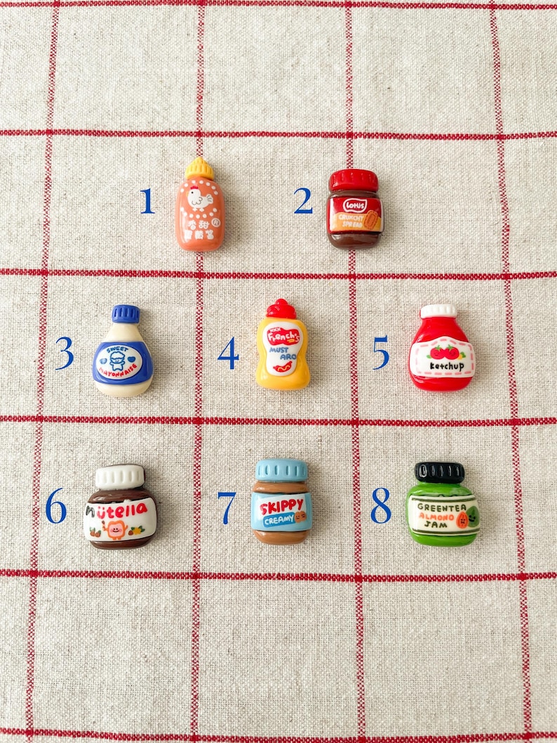 May include: Eight miniature ceramic jars and bottles with various food labels, including ketchup, mustard, peanut butter, jam, and Nutella. The jars are arranged in a grid pattern on a white and red checkered surface.