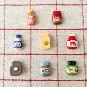 May include: A set of nine miniature food jar magnets, including ketchup, peanut butter, jam, and more. The magnets are colorful and feature cartoon-like illustrations.