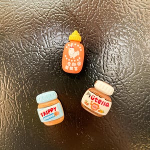 May include: Three miniature food jar magnets. A red and orange jar with a white chicken and the text "香辣鸡" in Chinese characters. A blue and brown jar with the text "SKIPPY CREAMY" in white. A brown jar with the text "NUTELLA" in white.