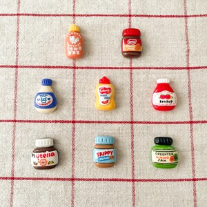May include: Nine miniature plastic jars and bottles with various food labels, including Nutella, Skippy, Green Tea Almond Jam, Sweet Strawberry, Ketchup,  French's Mustard, and a jar of Creamy Spread. The jars and bottles are arranged in a grid pattern on a white and red checkered background.