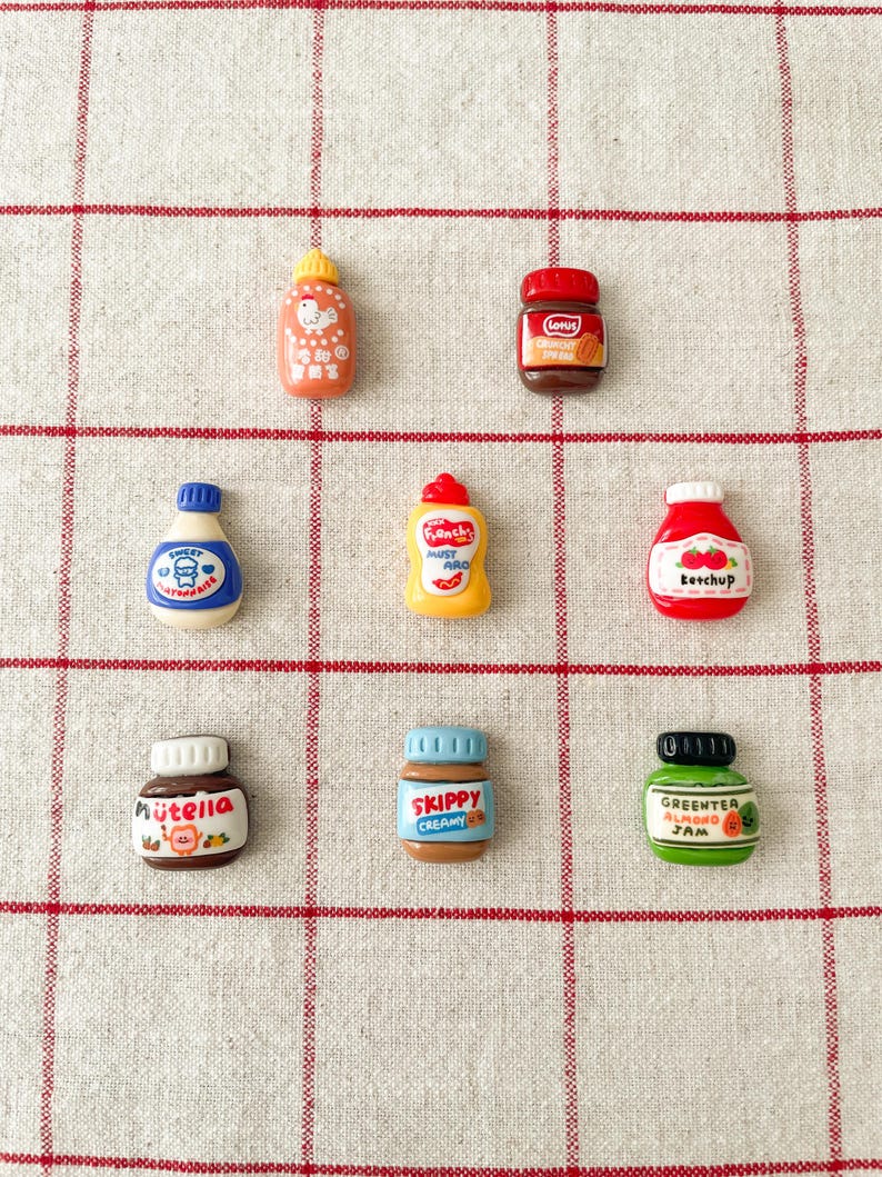 May include: Nine miniature plastic jars and bottles with various food labels, including Nutella, Skippy, Green Tea Almond Jam, Sweet Strawberry, Ketchup,  French's Mustard, and a jar of Creamy Spread. The jars and bottles are arranged in a grid pattern on a white and red checkered background.