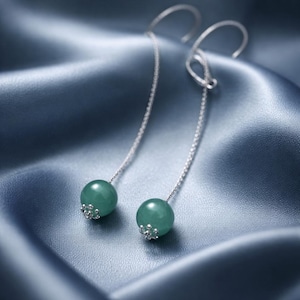 May include: A pair of elegant drop earrings. Each earring features a silver chain suspending a round, green gemstone bead. The beads are accented with silver floral-like caps. The earrings are displayed against a backdrop of blue fabric.