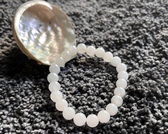 May include: A white beaded bracelet with round beads is displayed next to a pearlescent seashell. The bracelet is resting on a textured gray surface. The beads appear to be made of a translucent material.