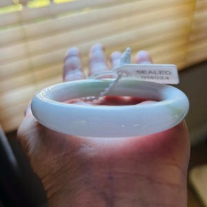 Könnte beinhalten: Ein weißes Jade-Armband mit grünem Farbton. Das Armband befindet sich auf einer Hand und hat ein Etikett mit der Aufschrift "SEALED 014024".