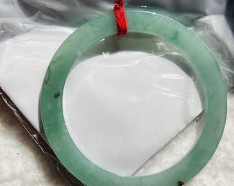 May include: A light green jade bangle bracelet with a polished finish. The circular bracelet is suspended by a red cord. The jade has natural variations in color and texture, with some darker green spots.