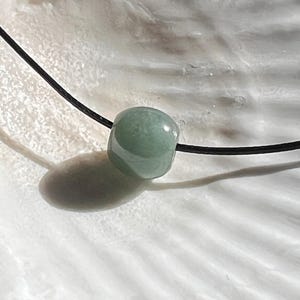 May include: A light green, rounded gemstone bead strung on a black cord. The bead is smooth and polished, with a subtle, translucent quality. The necklace is displayed against a white, textured background.