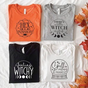 May include: Four different t-shirts with witchy designs. The first shirt is orange with black text that says "Hex the Patriarchy" with a moon and star design. The second shirt is gray with black text that says "There's a little witch in all of us" with a moon and star design. The third shirt is black with white text that says "Feeling Witchy" with a moon and star design. The fourth shirt is white with black text that says "Spell casting in progress" with a snow globe design.