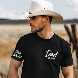 Może przedstawiać: Czarny T-shirt z białym tekstem, który brzmi "Dad Est. 2023". Lewy rękaw ma biały tekst, który brzmi "Olivia Rachel".