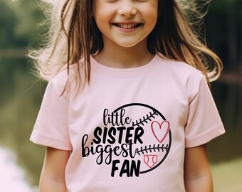 Może przedstawiać: Mała dziewczynka w różowej koszulce z grafiką baseballową i napisem "little sister biggest fan" w kolorze czarnym. Grafika baseballowa ma czerwone serce i czerwony szew baseballowy.