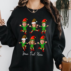 May include: Black sweatshirt with a design of six elves with different faces. The elves are wearing green and red outfits and have red hats. The text "Your Text Here" is printed below the elves.