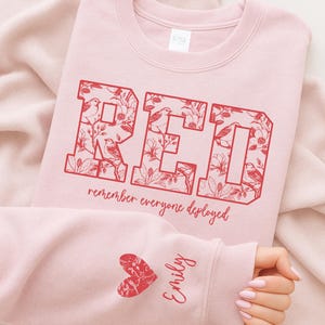 May include: A light pink crewneck sweatshirt featuring the red text RED in a block font filled with floral and bird illustrations. Below the text is the phrase remember everyone deployed. The sleeve features a red heart and the name Emily.