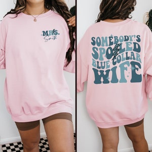May include: Pink sweatshirt with a blue and white graphic on the back that says "Somebody's Spoiled Blue Collar Wife" and a smaller blue graphic on the front that says "Mrs. Smith".