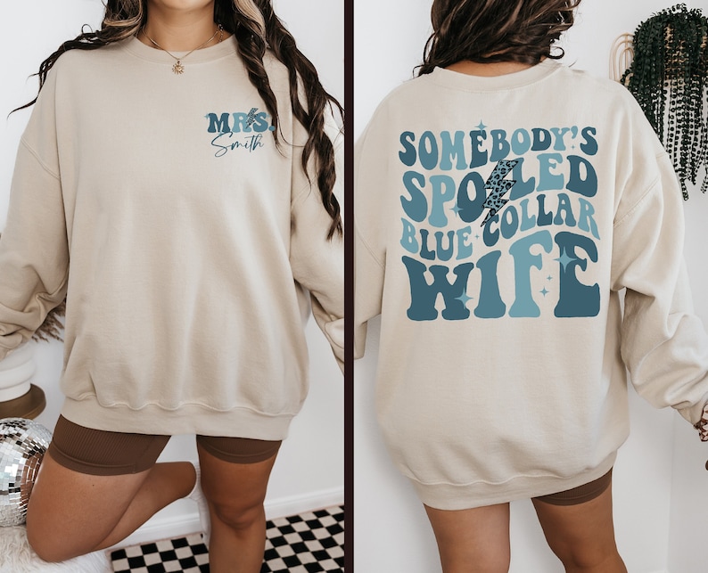 May include: A beige sweatshirt with the text "MRS. Smith" on the front and "SOMEBODY'S SPOILED BLUE COLLAR WIFE" on the back. The text is in a blue and white retro font with a lightning bolt graphic.