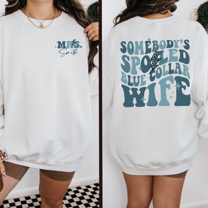 May include: White sweatshirt with blue and white text that says "Somebody's Spoiled Blue Collar Wife" with a lightning bolt graphic. The front of the sweatshirt has the text "Mrs. Smith" in blue.