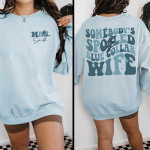 May include: Light blue sweatshirt with a retro-style graphic on the back that says "Somebody's Spoiled Blue Collar Wife" and a smaller graphic on the front that says "Mrs. Smith".