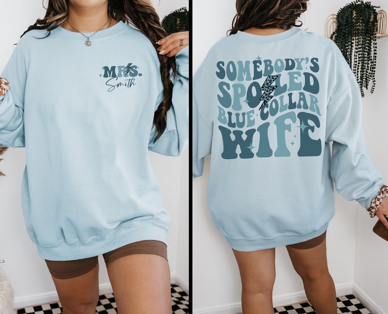 May include: Light blue sweatshirt with a retro-style graphic on the back that says "Somebody's Spoiled Blue Collar Wife" and a smaller graphic on the front that says "Mrs. Smith".
