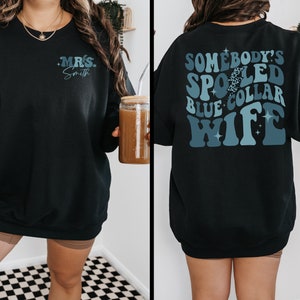 May include: Black sweatshirt with a blue and white graphic that says "Somebody's Spoiled Blue Collar Wife" on the back and "Mrs. Smith" on the front.