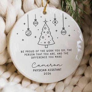 May include: A round white ceramic ornament featuring a black line drawing of a Christmas tree and hanging ornaments. The text reads: Be proud of the work you do, the person that you are, and the difference you make, Cameron, Physician Assistant, 2026.