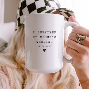May include: White ceramic mug with the text "I SURVIVED MY NIECE'S WEDDING 09.20.2025" in black, with a small heart symbol. The mug is held by a person wearing a black and white checkered hat and a pink sweater.