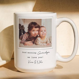 Peut inclure: Un mug en céramique blanche orné d'une photographie rectangulaire de deux enfants allongés. Sous l'image, le texte indique : Good morning Grandpa we love you! Keva & Nate. Le mug possède une anse incurvée et une finition lisse et brillante.