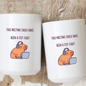 May include: Two white ceramic mugs with the text "THIS MEETING COULD HAVE BEEN A FIST FIGHT". Each mug features a cartoon capybara wearing headphones and using a laptop. The mugs have large white handles.
