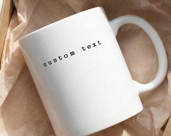 Op de afbeelding: Een witte keramische mok met de woorden "custom text" in het zwart gedrukt, in een bruine kartonnen doos met gekreukeld papier. De mok heeft een grote, gebogen handgreep.