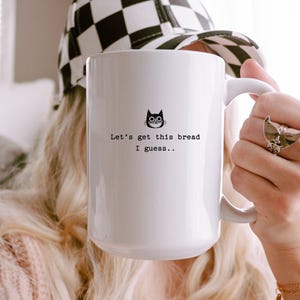 May include: White ceramic mug with a black cat illustration and the text "Let's get this bread I guess..". The mug is held by a person wearing a black and white checkered hat.