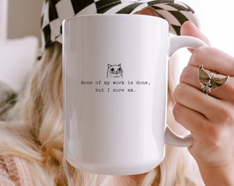 May include: White ceramic mug with a cat illustration and the text "None of my work is done, but I sure am." The mug has a large handle and is held by a person. A black and white checkered hat is visible in the background.