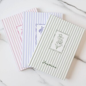 May include: Three notebooks with striped covers in pink, lavender, and green. Each notebook features a floral illustration within a rectangular frame. The green notebook is labeled "Jasmine" in cursive.