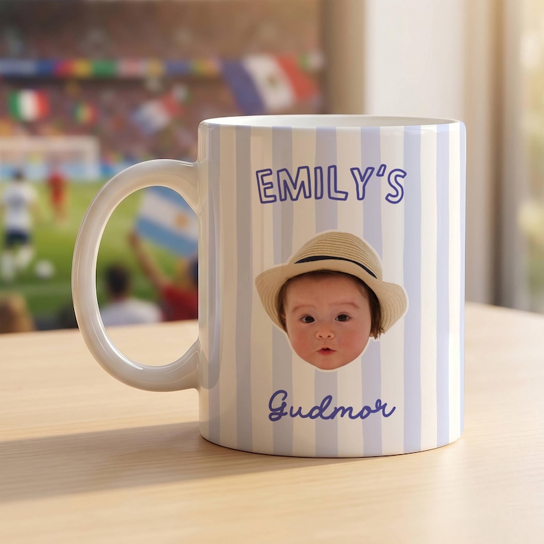 Puede incluir: Una taza de cer&aacute;mica blanca con rayas verticales de color azul claro. La taza muestra el texto EMILY'S en la parte superior y Gudmor en la parte inferior en azul. Centrada entre el texto hay una foto de un beb&eacute; con un sombrero de paja beige.