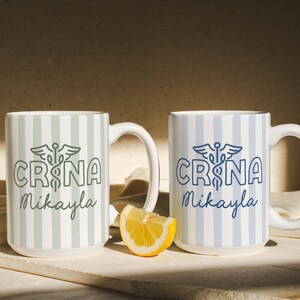Pu&ograve; includere: Due tazze in ceramica bianca con motivi a righe verticali. Una tazza ha strisce e testo verdi, l'altra ha strisce e testo blu. Entrambe le tazze presentano il testo "CRNA Mikayla" e un simbolo medico.