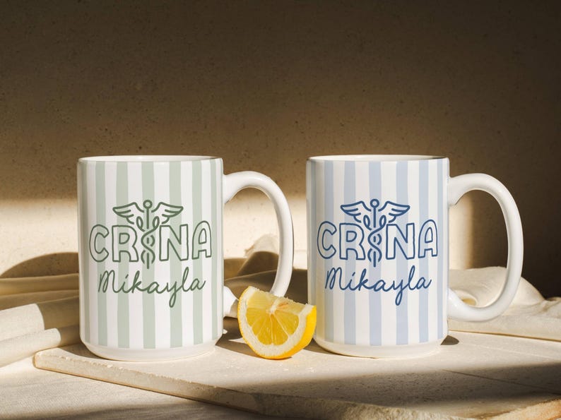 Pu&ograve; includere: Due tazze in ceramica bianca con motivi a righe verticali. Una tazza ha strisce e testo verdi, l'altra ha strisce e testo blu. Entrambe le tazze presentano il testo "CRNA Mikayla" e un simbolo medico.