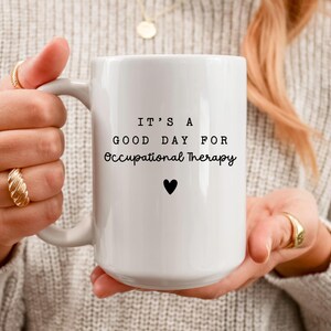 May include: White ceramic mug with the text "IT'S A GOOD DAY FOR Occupational Therapy" in black, with a small black heart. The mug is held by a person wearing gold rings and a sweater.