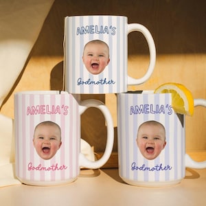 May include: Three white ceramic mugs with vertical striped patterns in pink and blue. Each mug features a baby's face and the text "AMELIA'S" and "Godmother". A lemon slice rests on the rim of one mug. The mugs have large, curved handles.