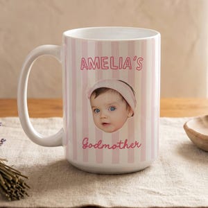 Puede incluir: Una taza de cer&aacute;mica blanca con un patr&oacute;n de rayas verticales en color rosa claro y blanco. La taza muestra una fotograf&iacute;a de un beb&eacute; con una diadema rosa, centrada entre el texto rosa AMELIA'S arriba y Godmother abajo.