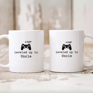 May include: Two white ceramic mugs with black handles. The mugs have a black video game controller graphic and the text "level up to Uncle" printed on them.
