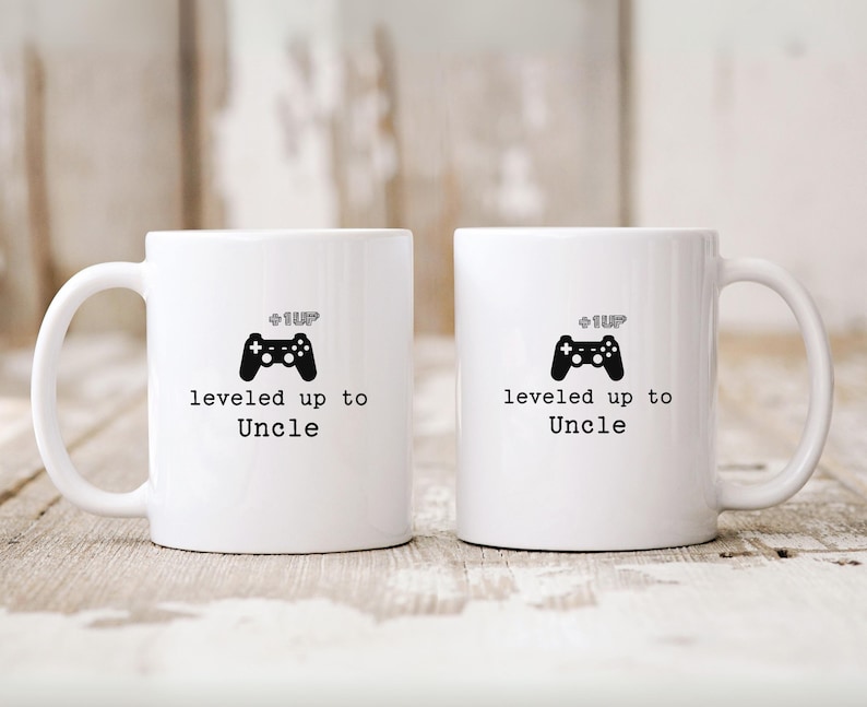 May include: Two white ceramic mugs with black handles. The mugs have a black video game controller graphic and the text "level up to Uncle" printed on them.