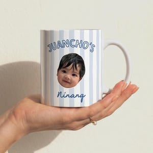Puede incluir: Una taza de cer&aacute;mica blanca con un patr&oacute;n de rayas verticales de color azul claro. La taza muestra una fotograf&iacute;a circular del rostro de un ni&ntilde;o peque&ntilde;o en el centro, con el texto JUANCHO'S impreso arriba y la palabra Ninang abajo en azul.