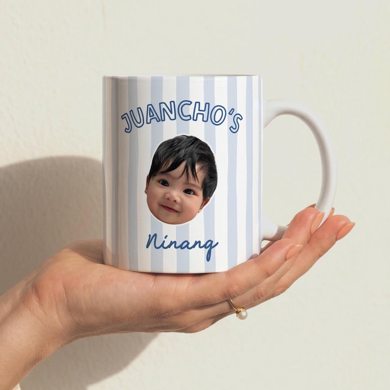 Puede incluir: Una taza de cer&aacute;mica blanca con un patr&oacute;n de rayas verticales de color azul claro. La taza muestra una fotograf&iacute;a circular del rostro de un ni&ntilde;o peque&ntilde;o en el centro, con el texto JUANCHO'S impreso arriba y la palabra Ninang abajo en azul.