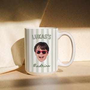 Puede incluir: Una taza de cer&aacute;mica blanca con un patr&oacute;n de rayas verticales en color verde claro y blanco. La taza muestra un retrato fotogr&aacute;fico de un ni&ntilde;o con gafas de sol rojas, con el texto LUKAS'S impreso arriba y la palabra Madrina debajo de la imagen.