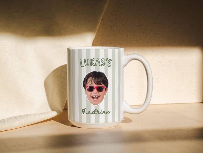 Puede incluir: Una taza de cer&aacute;mica blanca con un patr&oacute;n de rayas verticales en color verde claro y blanco. La taza muestra un retrato fotogr&aacute;fico de un ni&ntilde;o con gafas de sol rojas, con el texto LUKAS'S impreso arriba y la palabra Madrina debajo de la imagen.