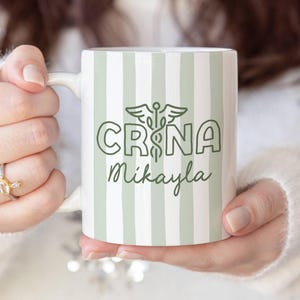 Pu&ograve; includere: Tazza in ceramica bianca con un motivo a righe verdi e bianche. La tazza presenta il testo "CRNA Mikayla" in verde, insieme a un simbolo di caduceo. La tazza &egrave; tenuta da una persona.