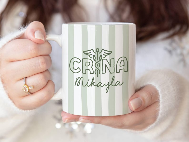 Pu&ograve; includere: Tazza in ceramica bianca con un motivo a righe verdi e bianche. La tazza presenta il testo "CRNA Mikayla" in verde, insieme a un simbolo di caduceo. La tazza &egrave; tenuta da una persona.