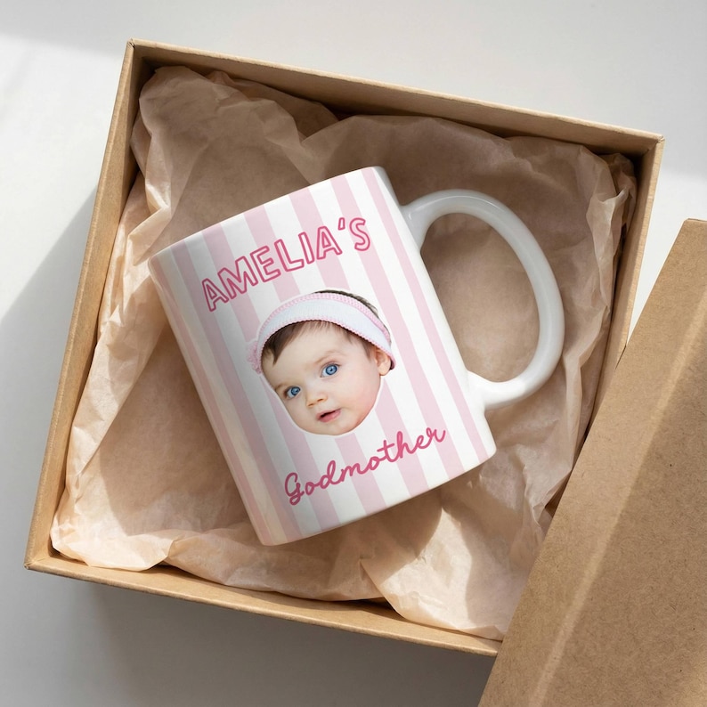 Puede incluir: Una taza de cer&aacute;mica blanca con un patr&oacute;n de rayas verticales de color rosa claro y blanco. La taza muestra una fotograf&iacute;a de un beb&eacute; con una diadema blanca, con el texto AMELIA'S impreso arriba y la palabra Godmother debajo de la imagen.
