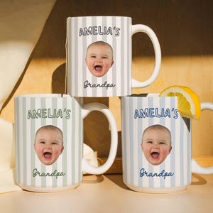 Peut inclure: Trois mugs en céramique blanche avec des motifs à rayures verticales grises, bleu clair et vertes. Chaque mug présente le visage d'un bébé et le texte "AMELIA'S Grandpa". Une tranche de citron repose sur un mug. Les mugs ont des anses blanches.