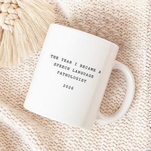 Op de afbeelding: Witte keramische mok met de tekst "THE YEAR I BECAME A SPEECH LANGUAGE PATHOLOGIST 2026". De mok staat op een cr&egrave;mekleurig gebreid oppervlak met een beige macram&eacute; decoratie.